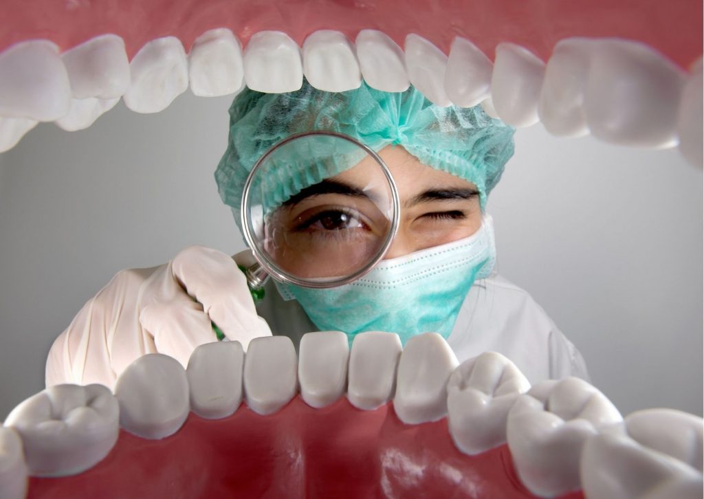 Mujer con gorro y guantes quirúrjicos con una lupa simula mirar el interior de la boca de una persona. Tema: ¿Cómo sé que tengo cáncer de boca?