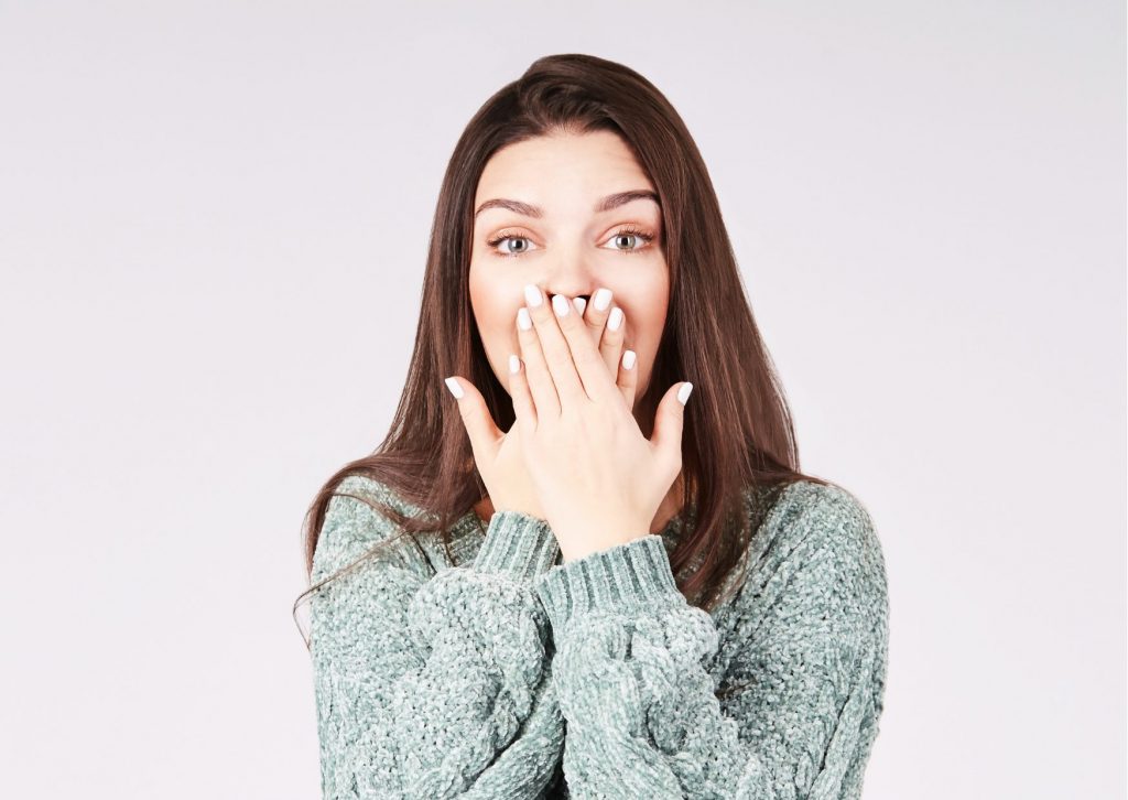 Mujer de cabello largo se tapa la boca con las dos manos para que no le vean los dientes. Fondo claro. Tema: diente negro después de una endodoncia