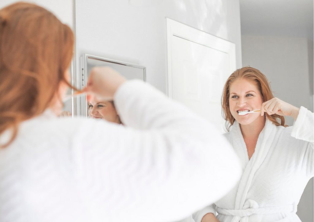 Mujer colorina cepilla sus dientes frente al espejo del baño vistiendo una bata de levantar color blanco. Mitos sobre la higiene dental