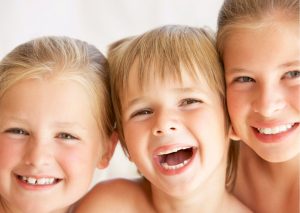 Dos niñas y un niño, rubios, de diferentes edades sonríen a la cámara. Tema: higiene bucal en niños, nota del Blog de Clínica Everest