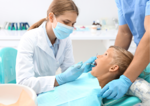 Mujer dentista rubia con mascarilla, examina la boca de un paciente infantil rubio en la consulta dental. Tema: Avulsión dental en niños de Blog Clínica Everest