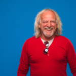 Hombre blanco de la tercera edad con cabello canoso y semi largo, con suéter de color rojo, ríe feliz a la cámara. Fondo azul. Tema: Prótesis dentales con ganchos transparentes para blog de Everest Life