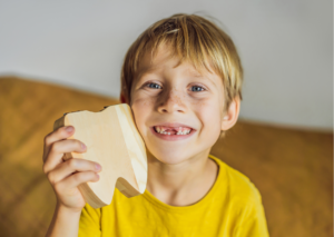 Niño rubio con pecas sonríe mostrando que le faltan algunos diente superiores, mientras sostiene un diente de madera de gran tamaño junto a su cara. tema: cambio de dientes de blog Everest Life