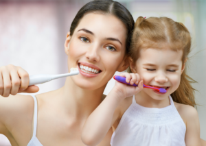Mujer de cabello oscuro junto a una niña pequeña de cabello claro lavan sus dientes frente al espejo. tema Fluorosis infantil para blog Everest Life