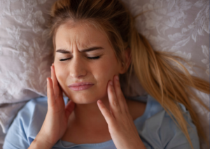 Mujer rubia de cabello largo acostada en su cama se toca con gesto de dolor la mandíbula. Tema: el bruxismo y sus consecuencias para Everest Life.