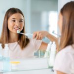 Adolescente mujer se lava los dientes frente al espejo del baño utilizando un cepillo dental eléctrico.