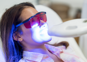 Mujer sentada en sillón dental con gafas de odontología tiene una luz azul en la mitad inferior de su casa. La paciente se somete a tratamiento de Blanqueamiento Dental Profesional