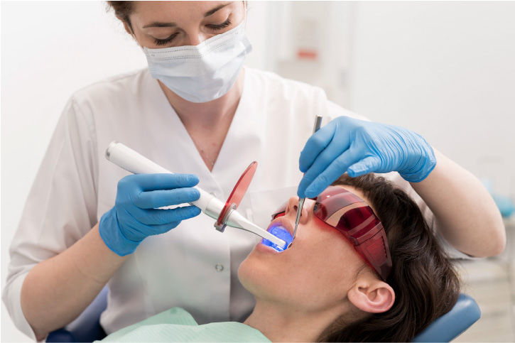 Paciente recostado en un sillón dental, siendo tratado por una odontóloga con una luz infrarroja y gafas, preparado para realizar su higiene o limpieza dental.